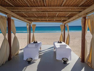 Beach massage beds under a wooden canopy with curtains by the sea.