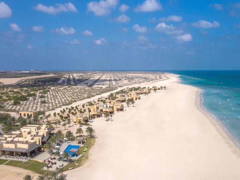 Aerial view of a large beach resort with white sand, blue pool, palm trees, and clear ocean water.