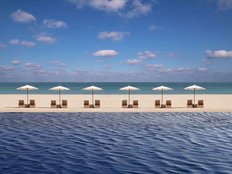 Row of six sunbeds with white umbrellas by blue pool and sandy beach under blue sky.