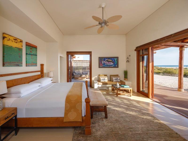 Bright hotel room with large bed, artwork, ceiling fan, and open doors to outdoor terrace and sea view.