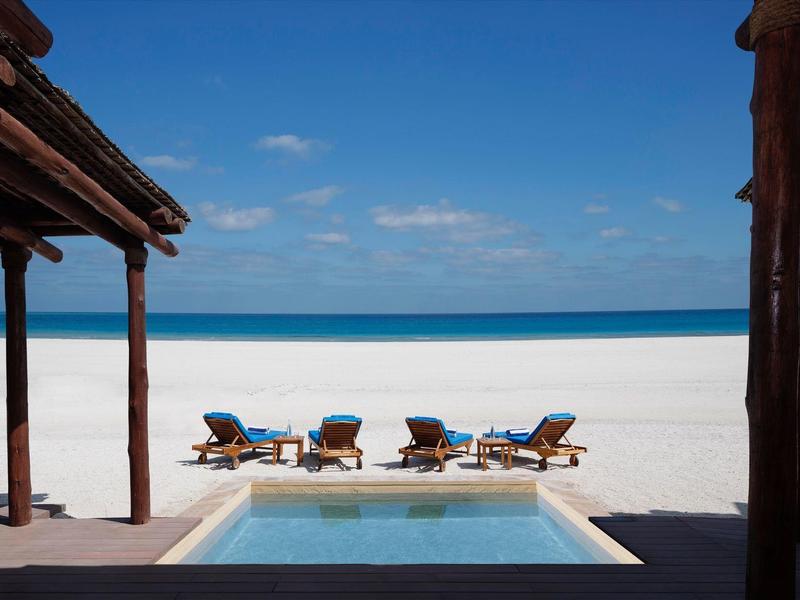 Four lounge chairs by a pool overlooking a white sandy beach and blue sky.