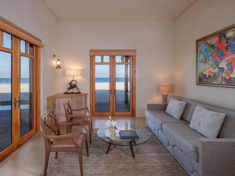 Cozy living room with gray couch, wooden chairs, glass table, and ocean view through wooden doors.