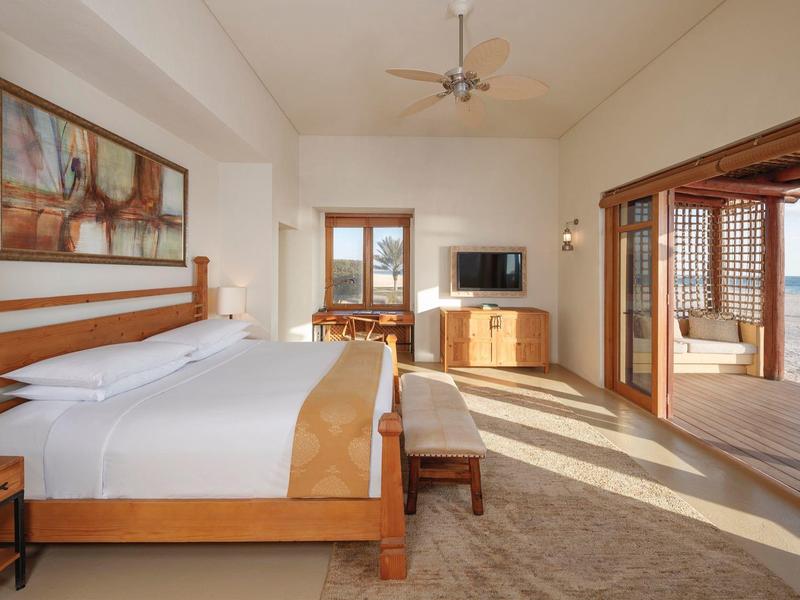 Spacious hotel room with wooden bed, bench, TV, and sliding door opening to sunny ocean view.