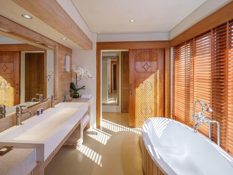 Bright hotel bathroom with freestanding tub, double vanity, wooden blinds, and floral decor.
