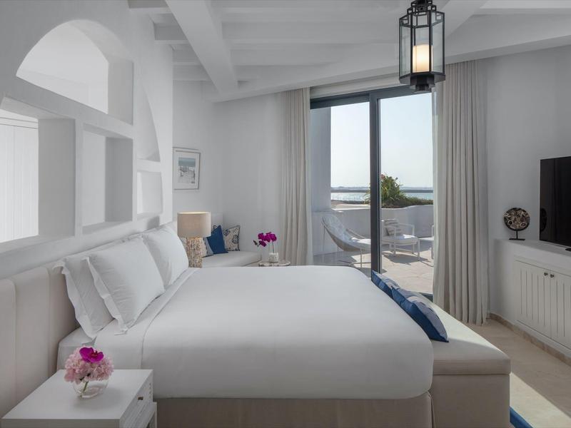 Bright hotel room with large bed, TV, and terrace overlooking the sea.