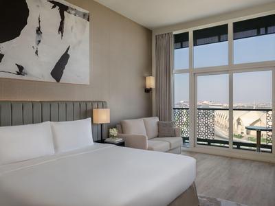 Modern eingerichtetes Hotelzimmer mit großem Bett, Sessel und bodentiefen Fenstern mit Stadtblick.