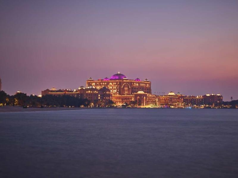 Illuminated luxury hotel by the water at sunset with pink and purple sky.