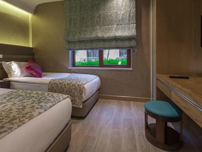 Hotelzimmer mit zwei Einzelbetten, Fenster mit Jalousie, Holzboden und Schreibtisch mit Hocker.