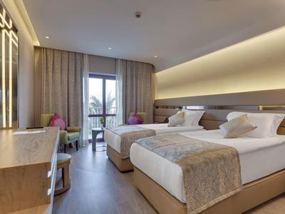 Modernes Doppelzimmer mit zwei Betten, Fenstertür zum Balkon, hellem Holzboden und sanfter Beleuchtung.