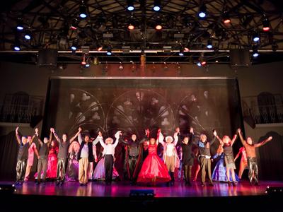 Grupa osób w kolorowych kostiumach kłaniających się na scenie pod ciepłym oświetleniem.