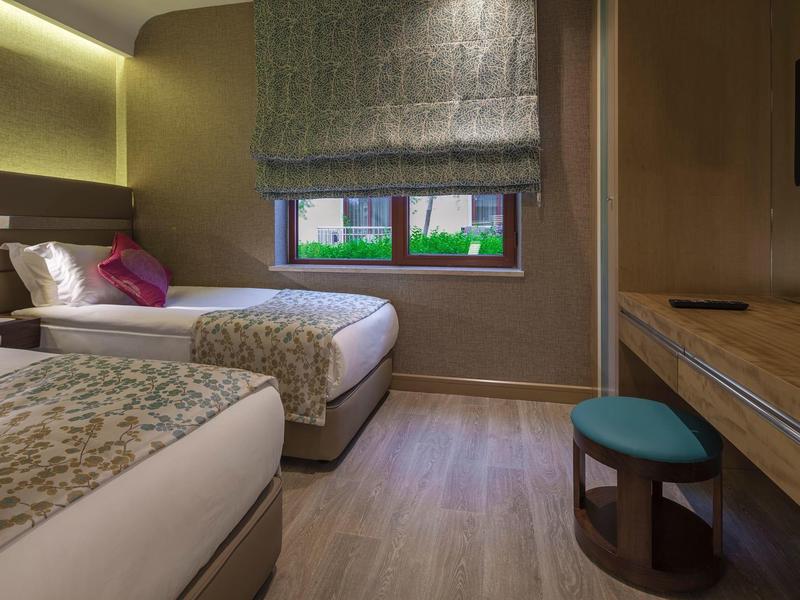 Hotelzimmer mit zwei Einzelbetten, Fenster mit Jalousie, Holzboden und Schreibtisch mit Hocker.