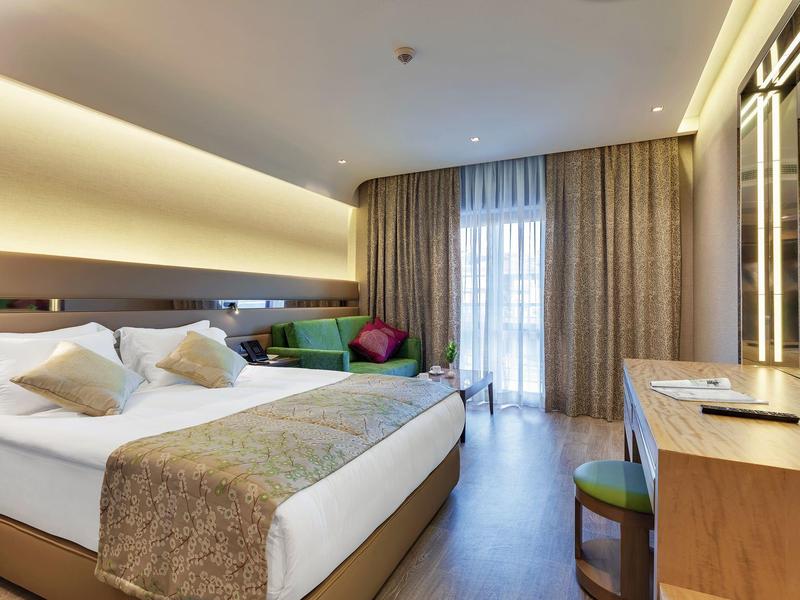 Modernes Hotelzimmer mit großem Bett, Schreibtisch, Fernseher und bodentiefem Fenster mit Vorhängen.