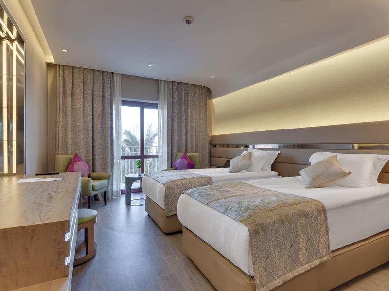 Modernes Doppelzimmer mit zwei Betten, Fenstertür zum Balkon, hellem Holzboden und sanfter Beleuchtung.