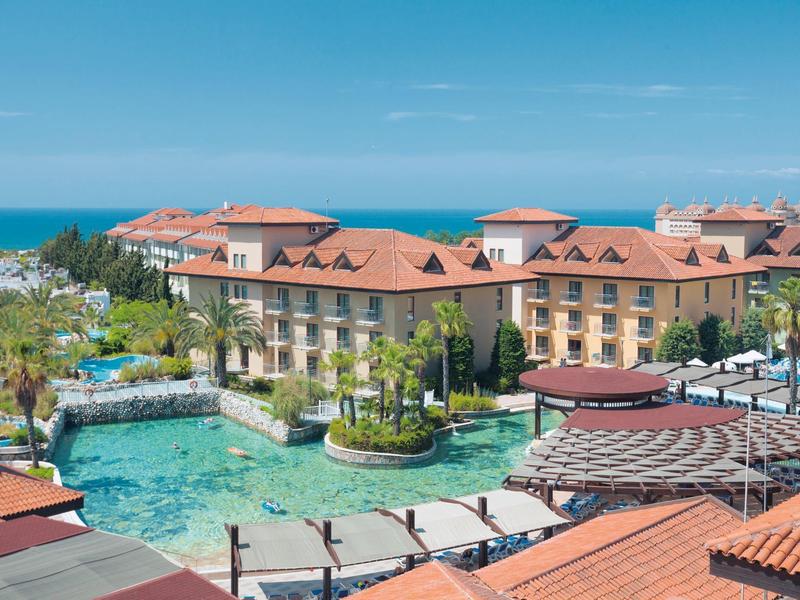 Großes Hotel mit rotdachigen Gebäuden, Pool, Palmen und Blick aufs Meer bei klarem Himmel.