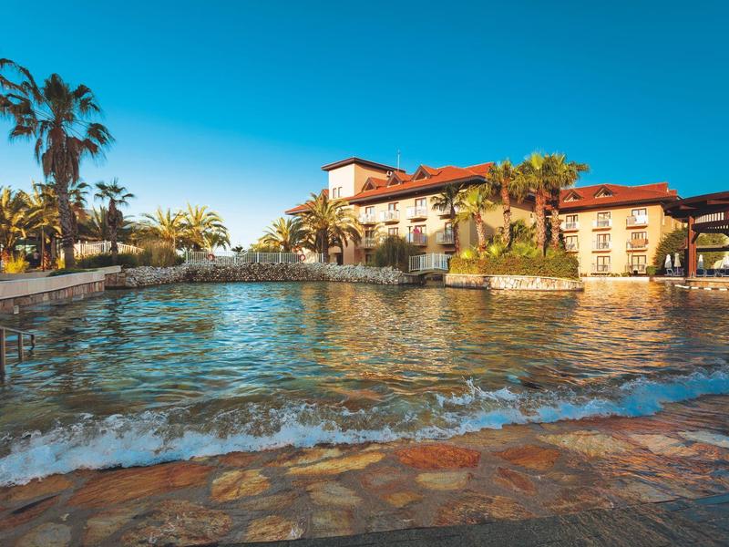 Großer Hotelpool mit klarem Wasser, umgeben von Palmen und mehrstöckigem Gebäude bei blauem Himmel.