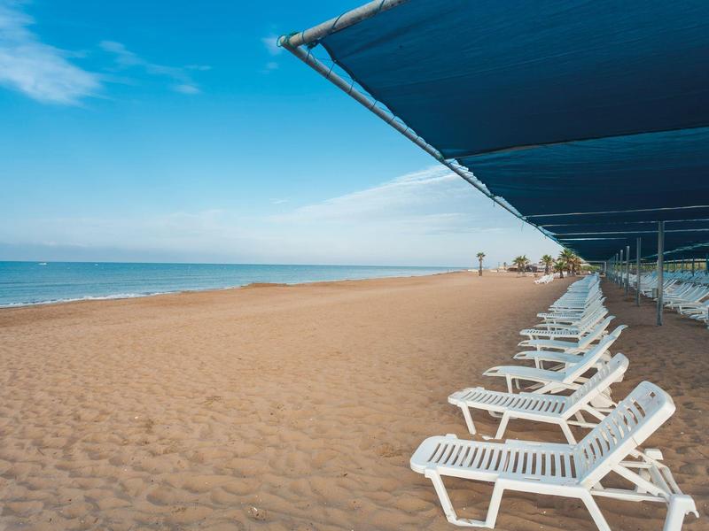 Leerer Sandstrand mit weißen Liegestühlen unter blauer Markise und klarem Himmel.