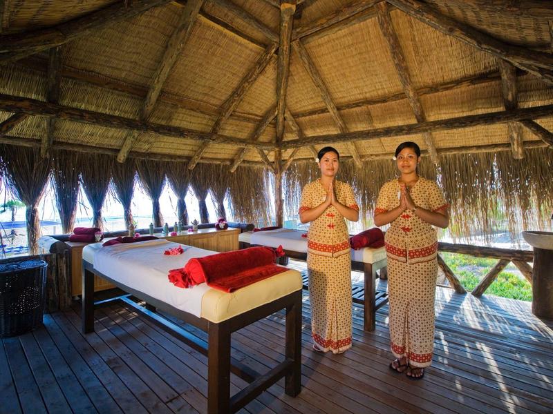 Zwei Frauen im traditionellen Gewand begrüßen Gäste in einer hölzernen Massagehütte mit Liegen.