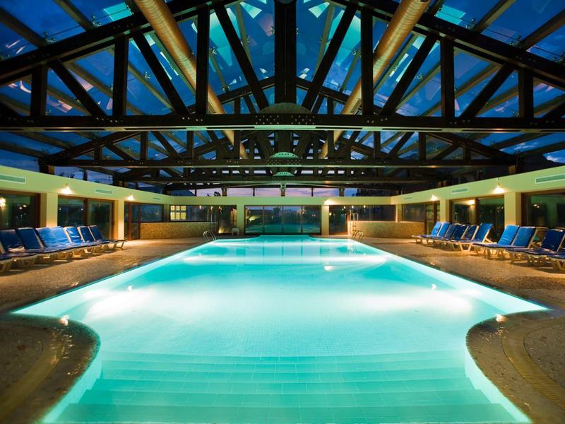 Großes Innenhallenbad mit blau beleuchtetem Wasser, Holzdeckenbalken und Liegen an den Seiten.