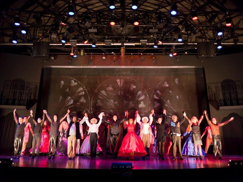 Bühnenensemble in farbenprächtigen Kostümen verbeugt sich vor Publikum im Theater mit Lichtshow.