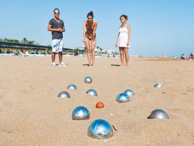 Drei Personen spielen Boule mit silbernen Kugeln und einer orangenen Zielkugel am sandigen Strand.