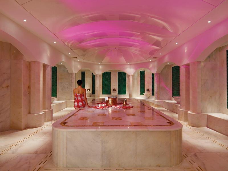 Luxe spakamer met roze verlicht plafond en centraal bubbelbad omringd door marmeren zuilen.