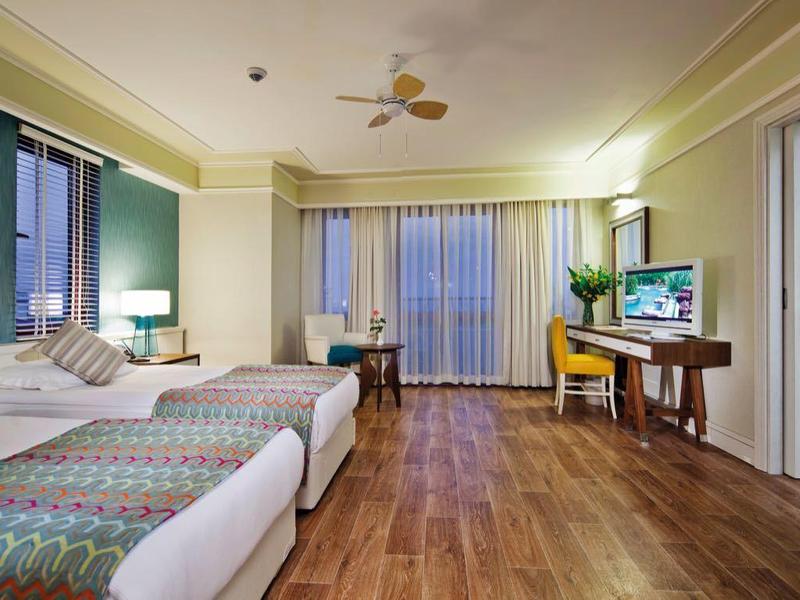 Ruime hotelkamer met twee bedden, houten vloer, bureau, tv, plafondventilator en grote ramen met gordijnen.