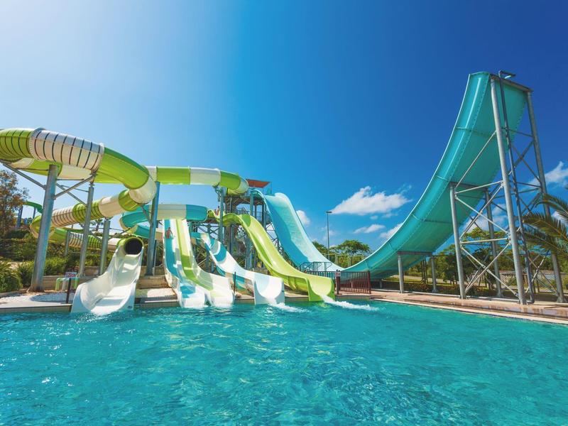 Gekleurde glijbanen leiden in een groot buitenzwembad onder een heldere blauwe lucht.