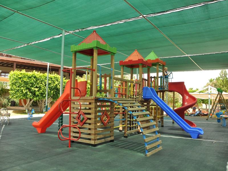 Buiten speelplaats met glijbanen en klimrekken onder groene schaduwnetten.