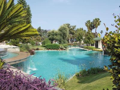 Großer blauer Pool umgeben von grünen Pflanzen, Palmen und lila Blumen unter blauem Himmel.