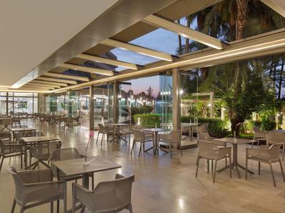 Moderner Außenbereich mit Glasdach, grauen Stühlen und Tischen, umgeben von Bäumen und Abendlicht.