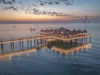 Ein Resort auf Stelzen über ruhigem Wasser bei Sonnenuntergang mit Steg und Booten im Hintergrund.