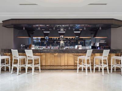 Bar mit hellem Holz, neun weißen Barhockern und dunklem Deckenaufbau, weißer Boden.