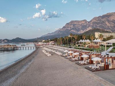 Strand mit Liegestühlen und Sonnenschirmen vor Hotels und Bergen im Hintergrund.