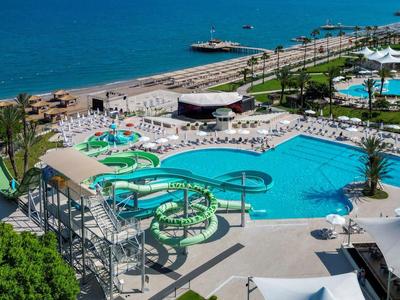 Großer Pool mit Wasserrutschen und Liegestühlen, umgeben von grünen Pflanzen und Meer im Hintergrund.