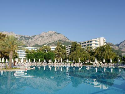 Großer Pool mit blauer Wasserfläche, Liegestühlen, Palmen und Hotelgebäuden vor Bergen.