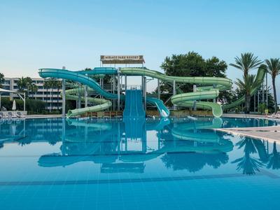 Wasserpark mit drei großen Rutschen über einem klaren blauen Pool, Palmen im Hintergrund.
