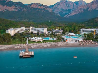 Küstenresort mit weißem Sandstrand, Liegen, Poolanlagen und Bergen im Hintergrund unter blauem Himmel.