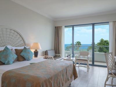 Helles Hotelzimmer mit großem Bett, zwei Nachttischen, Schreibtisch und Balkon mit Meerblick.