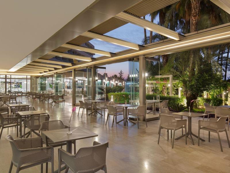 Moderner Außenbereich mit Glasdach, grauen Stühlen und Tischen, umgeben von Bäumen und Abendlicht.