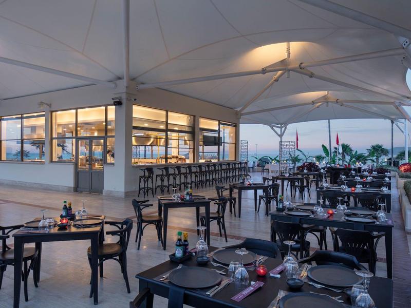 Außerhalb eines modernen Restaurants mit weißem Sonnendach und schwarzen Stühlen auf Terrassenboden.