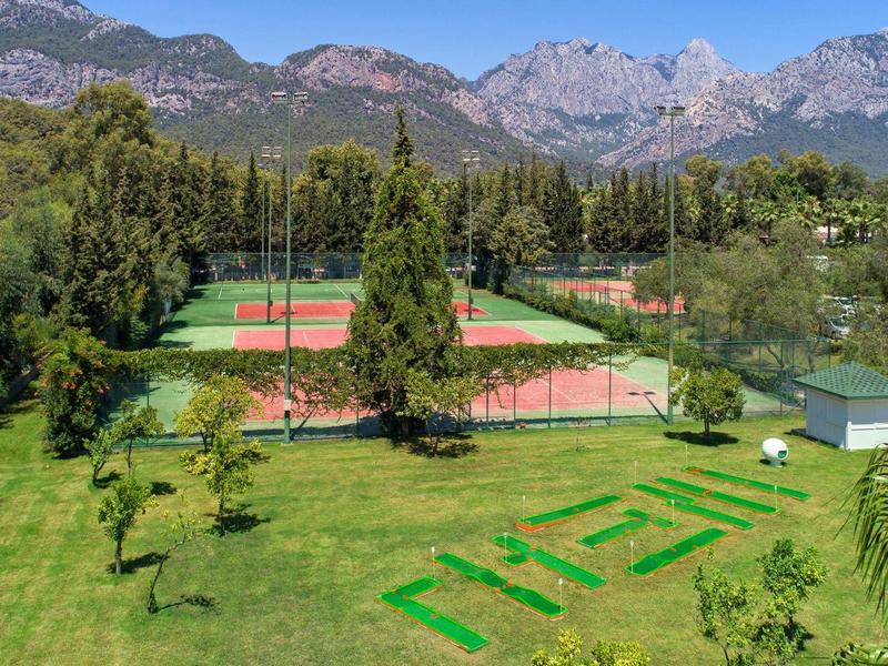 Grüne Wiese mit Tennisplätzen, Bäumen und Bergen im Hintergrund unter blauem Himmel.
