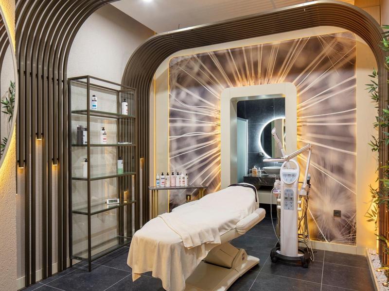 Moderner Behandlungsraum mit Praxisliege, Regal, dekorativer Wandbeleuchtung und Pflanzen im Raum.