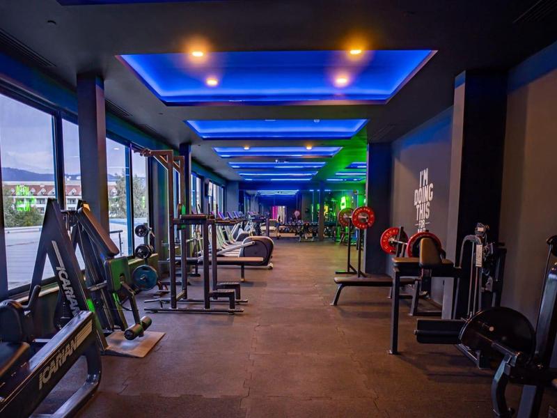 Moderner Fitnessraum mit großen Fenstern, blauer Deckenbeleuchtung und verschiedenen Trainingsgeräten.