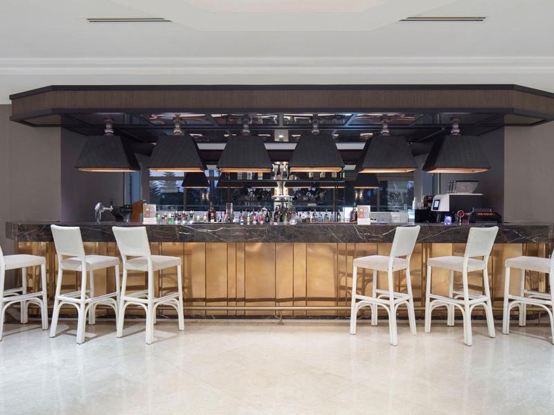 Bar mit hellem Holz, neun weißen Barhockern und dunklem Deckenaufbau, weißer Boden.