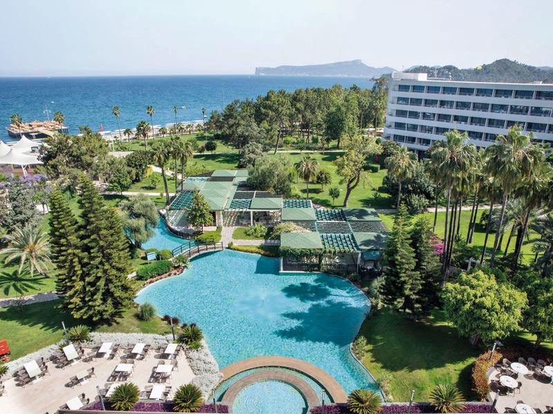 Großer Hotelpool umgeben von Palmen und Liegestühlen, Blick auf Meer und bewaldeten Hügel im Hintergrund.