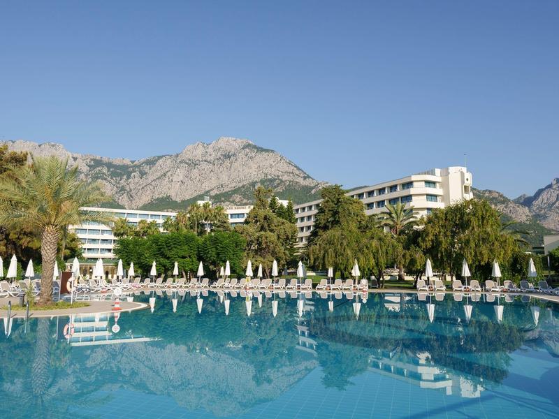Großer Pool mit blauer Wasserfläche, Liegestühlen, Palmen und Hotelgebäuden vor Bergen.