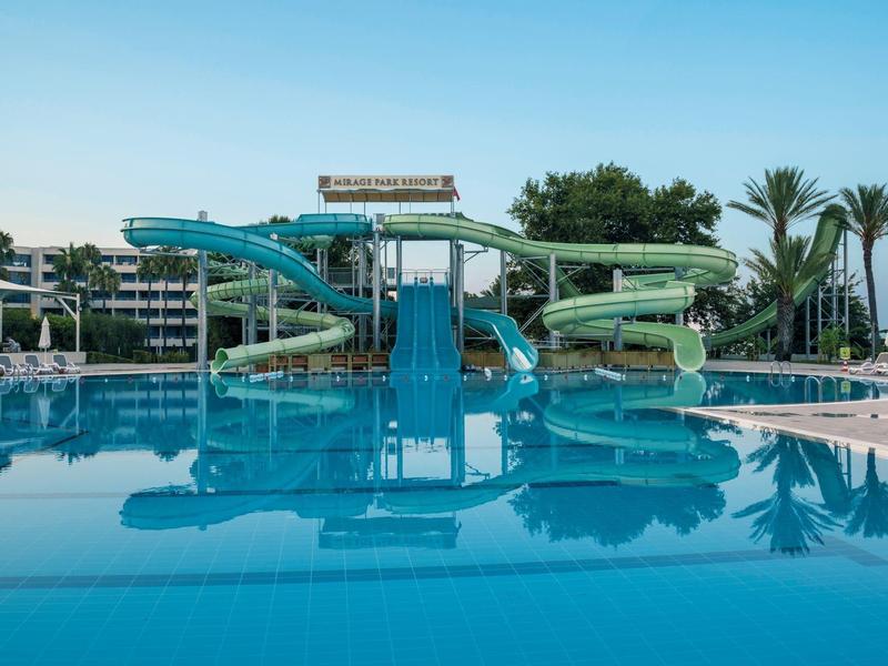 Wasserpark mit drei großen Rutschen über einem klaren blauen Pool, Palmen im Hintergrund.