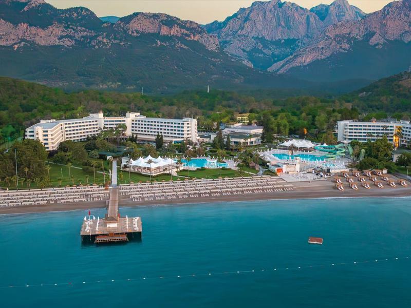 Küstenresort mit weißem Sandstrand, Liegen, Poolanlagen und Bergen im Hintergrund unter blauem Himmel.
