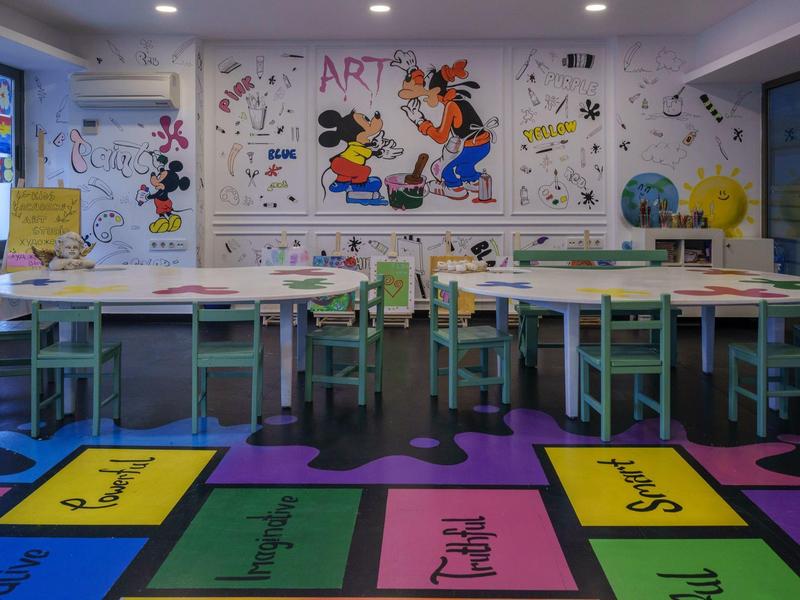 Bunter Klassenraum mit gemalten Figuren, kleinen Tischen, Stühlen und lehrreichem Bodenmuster.