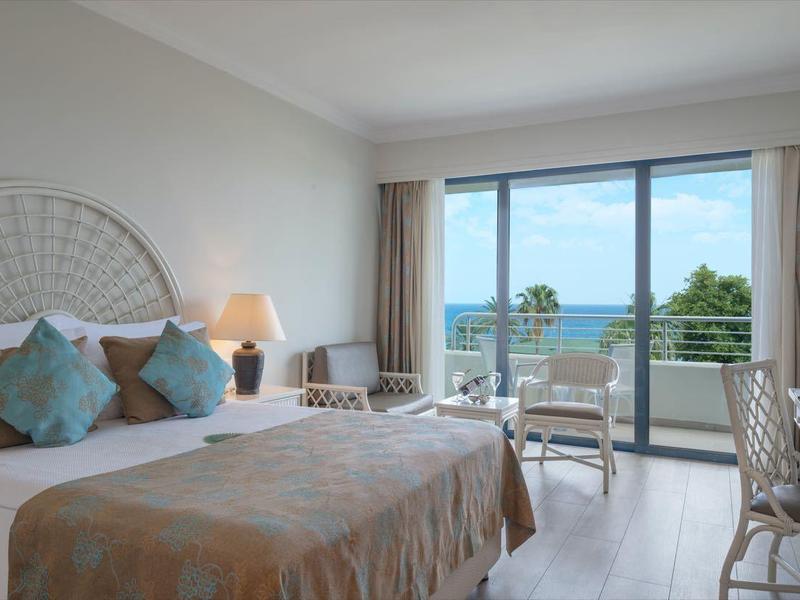 Helles Hotelzimmer mit großem Bett, zwei Nachttischen, Schreibtisch und Balkon mit Meerblick.